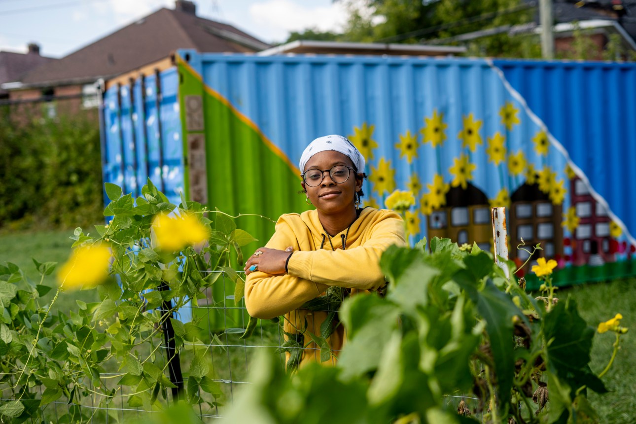A Black personwoman wearing a white headscarf, glasses and yellow hoodie smiles and leans on a garden fence near a blue shipping container with a mural of a neighborhood and sunflowers.